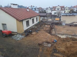 Plac budowy BCU, na placu maszyny budowlane pracownicy wykonujące prace ziemne. Widać stary budynek spawalni